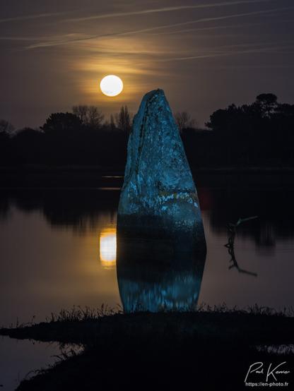 Photo en couleur en format vertical : le sujet principal est un menhir cerné par la marée haute en début de nuit, alors que la pleine lune s'est levée juste derrière le mégalithe et se reflète sur la surface tranquille de la ria envahie par la mer. Le menhir, les arbres de la rive opposée, ainsi qu'une branche plantée dans la vase à droite du mégalithe, se reflètent également à la surface de l'eau.