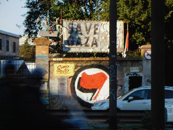 graffiti  of the anti-fascist symbol  (two flags,  a red one and a black one behind,  inside of a circle),  and the text  "SAVE  GAZA"  on a banner  above ,  at the entrance of the social center  'ex-SNIA'  in Rome