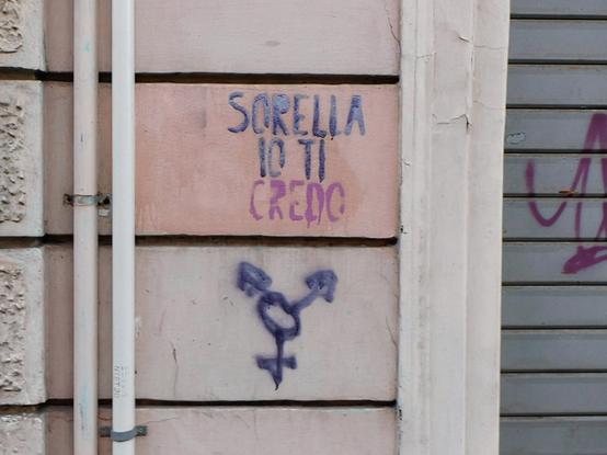 purple and pink  stencil text  on a street wall ,  reading "SORELLA  IO TI  CREDO"  (italian for 'i believe you, sister') ;   below it ,   trans symbol graffiti