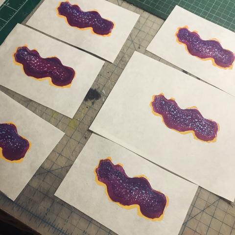 A collection of 6 tiny prints laid out on cutting mats of fuchsia flatworms on a white background. The animals are an amorphous wiggly oval shape, fuchsia with pale blue dots in the centre, a ring of fuchsia without dots and some yellow orange on its circumference.