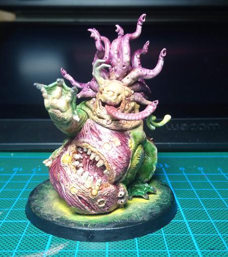 Frontview of a Beast of Nurgle Miniature. It raises its right flipper and a long, slobbering tongue is peeking out of its mouth. It is painted in purple and greenish/yellow tones.