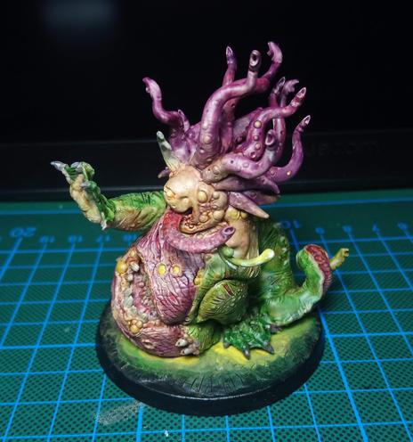 Sideview of a Beast of Nurgle Miniature. It raises its right flipper and a long, slobbering tongue is peeking out of its mouth. It is painted in purple and greenish/yellow tones.