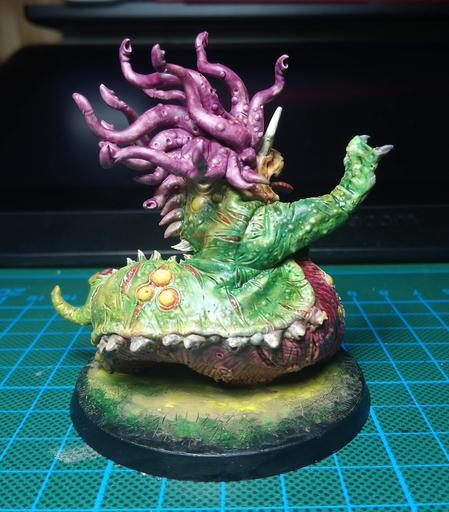 Sideview/backview of a Beast of Nurgle Miniature. It raises its right flipper and a long, slobbering tongue is peeking out of its mouth. It is painted in purple and greenish/yellow tones.