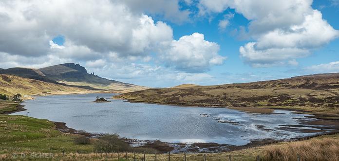 Panoramic view of Loch Leathan with a small stony island in the middle. 
The calm waters near the shore reflect a magnificent blue sky dotted with fluffy clouds. 
In the distance, the basalt pinnacle Old Man of Storr stands out against the horizon.