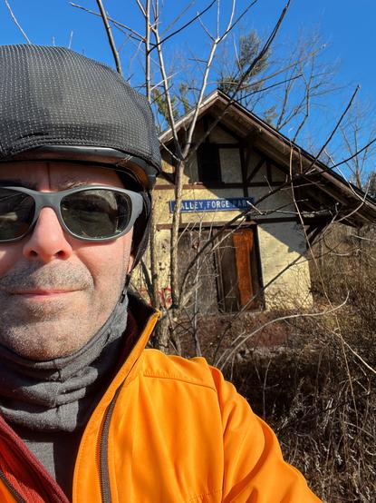 A selfie of the author in front of the abandoned and decrepit Valley Forge Park rail station.  I am on the left of the picture, looking fully into the camera, wearing a bicycle helmet (with a wind cover on top), dark sunglasses, as much of a smile as I ever muster in selfies, a few days' worth of beard stubble. There is a gray gaiter around my neck and a bright yellow jacket below. Behind me is the gable end of the abandoned Valley Forge Park railroad station, a half-timber building with bare painted brick on the first story and stucco above. There are large doors that appear to be on trolley hangers; above the doors is an old blue highway-style sign that reads "Valley Forge Park."
