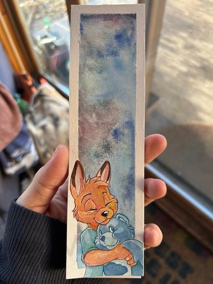 A hand holding a painted bookmark depicting a Joey cuddling a smiling Grumpy bear care bear plush. The background is a wash of blue watercolor.