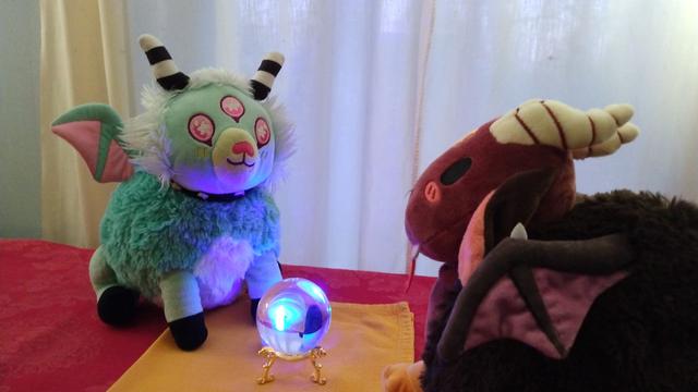 Two plushie demons -- one is light-blue green and pastel pink, with horns and wings. The other is red and brown, also with horns and wings. The pastel plush is gazing into a glowing crystal ball, while the red-brown one looks on.