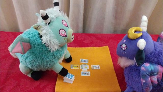 Two plushies -- one a pink and light blue green sheep-like figure, with striped horns and pink and blue wings, reading tarot cards, while a pink a purple sheep-like figure looks on.