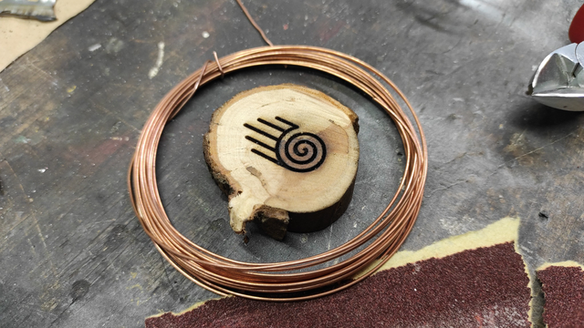 First photo illustrating the inlay process of a wooden piece resembling a branch slice, laser-engraved with a straightforward symbol—a spiral. On one side of the spiral, five parallel lines emerge, yet to be inlaid. A roll of copper wire surrounds the artwork.