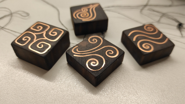Four additional pieces of wood, skillfully inlaid with copper, now complete and leveled. Unlike raw branch slices, these wooden pieces are uniform, low blocks with a square base. The inlays showcase imaginative spiral patterns—one resembling ocean waves, another resembling a drawn representation of a gust of wind, the third reminiscent of an Egyptian four-armed design, and the fourth bearing a slight resemblance to the Jitsi logo. After consideration, it may be four symbols of the air, fire, earth and water elements.