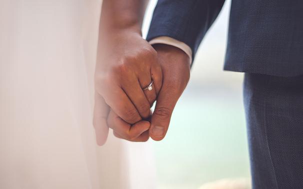 photo of two white hands holding each other with a wedding ring on one. In the background is part of a white dress on one side and a dark suit on the other
