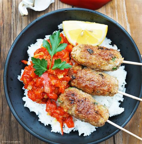 Holzhintergrund mit Maserung. Eine schwarze Schale mit Reis und Ajvar und Cevapcici aus Dinkelbulgur, die auf Spießen stecken. Die Geschichte jetzt auch als Audio.