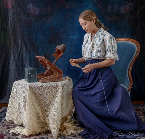 A woman wearing a floral blouse with a long blue skirt sitting in a blue victorian chair. She is holding a photo that can be used in a stereoscope. The stereoscope and a blue Kodak box camea are on a table to her left.