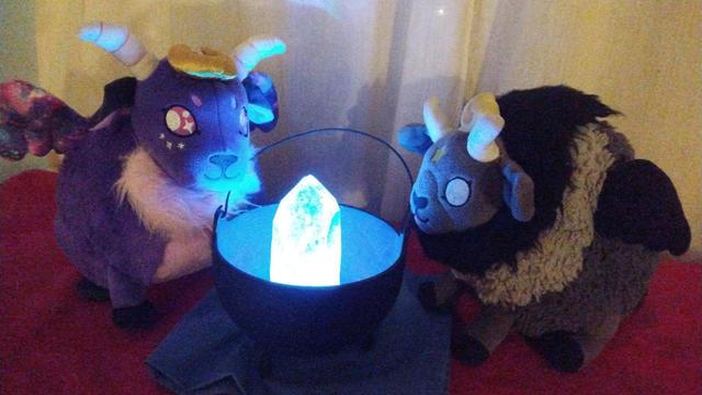 Two plushies, one a purple and pink sheep-like plushie, the other a black and grey goat-like plushie, gazing at a glowing crystal in a cauldron.
