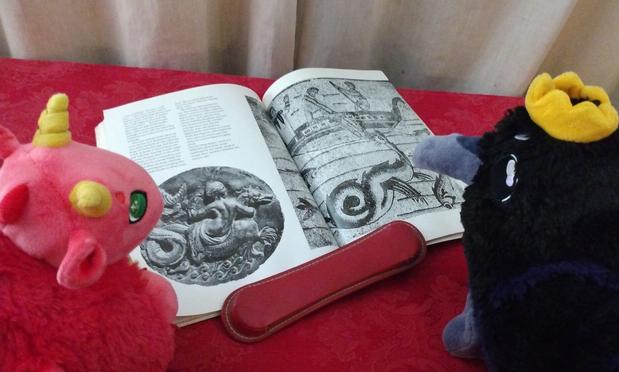 Two plushies looking at an open book. One is a red dragon plush, the other a raven plush wearing a crown. In the book are images of dragons.