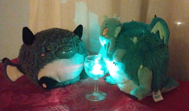 Two plushies -- one is a smiling grey and white bat -- the other a light green creature with tentacles and wings. They are looking at a crystal chalice with a glowing blue-green entity.
