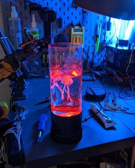 Somewhat messy desk with a partially filled aquarium and 2 (fake) jellyfish. Aside from the fact that it's currently wired into a cobbled-together power supply, there's no indication that the aquarium has been tampered with.
