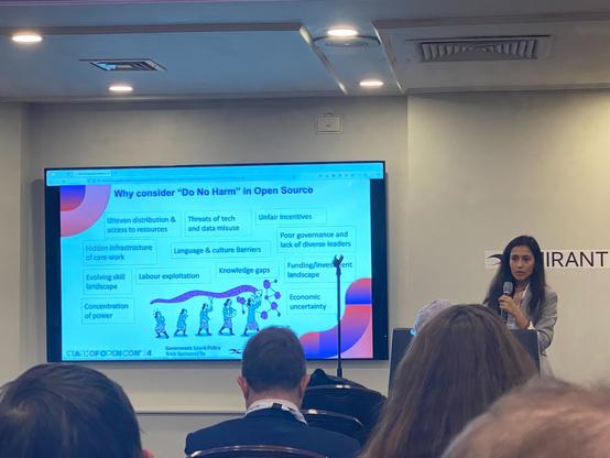 Malvika is standing in front of a room (wearing a grey blazer), holding a microphone and looking towards a screen that has the text: "'Why consider "Do No Harm" in open source. The answers include points from "uneven distribution & access to resources, hidden infrastructure of care work, evolving skill landscape, concentration of power, labour exploitation, language & cultural barriers, threats of tech and data misuse, language & culture barriers, knowledge gaps, unfair incentives, poor governance and lack of diverse leaders, funding/investment landscape, economic uncertainty"