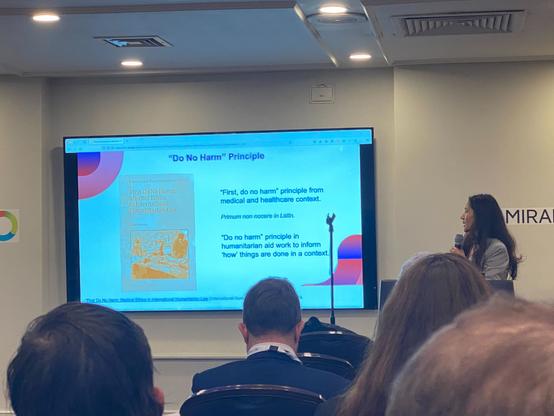 Malvika is standing in front of a room (wearing a grey blazer), holding a microphone and looking towards a screen that has the text: "'First, do no harm' principle from medical and healthcare context. 'Do no harm' principle in humanitarian aid work to inform 'how' things are done in a context."