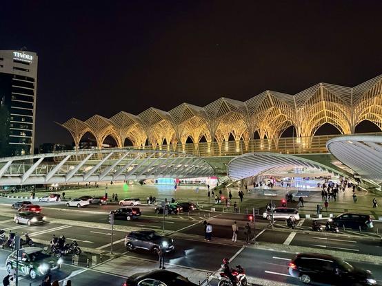 Das foto zeigt das beleuchtete Dach und den beleuchteten Vorplatz des Lissabon Oriente Bahnhofs bei Nacht. Links das beleuchtete Hotel Tivoli. Im Vordergrund dei Hauptverkehrsstraße mit bedeuteten Autos.