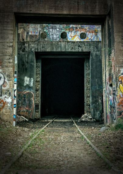 A photo taken in front of an open entrance into an old abandoned military train depot. It's a concrete structure with tags and graffiti, and in the middle a large gaping black hole where the train would roll in. The gate that is almost completely open consists of rectangular metal mesh sections filled with rocks.