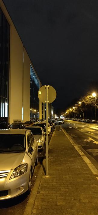 Ein schmaler Gehsteig der gesäumt ist von Parkstreifen und Autostraße, endet in 20m in einem weiteren Parkstreifen.