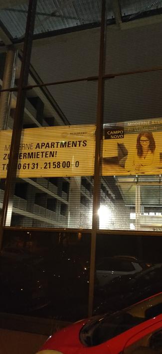 Reklame hinter einer gläsernen Lärmschutzwand: Moderne Apartments zu vermieten. Mit einer Businessfrau die in ihrem Wohnbüro lächelt. Davor noch ein dritter Bonus-Parkstreifen
