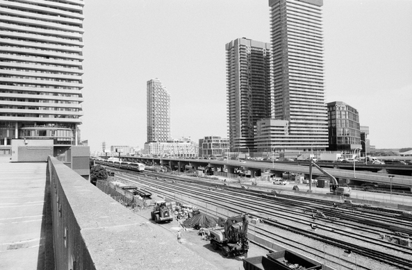 A black and white view from atop a parking garage of the former industrial harbour lands of downtown Toronto. The rail corridor and the elevated Gardiner Expressway are in full view with a GO train moving on the tracks. Several condo towers now loom and, in the distance, the last silos at this end of the harbour still stand.