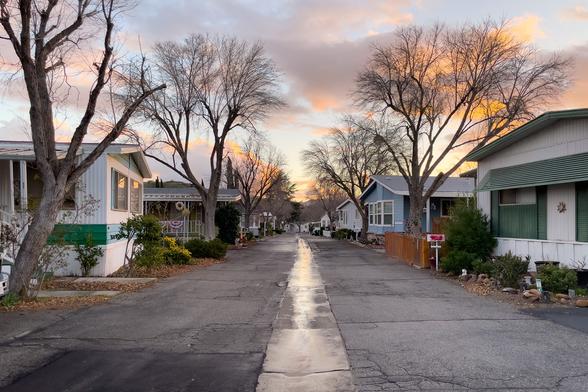 Glow of sunset cast through the clouds and reflected on a wet street in a trailer park. Trees and trailer homes frame the street.