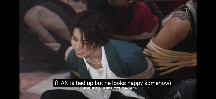 Screenshot of Han Jisung tied up with ropes around his chest and arms. Captioned "(HAN is tied up but he looks happy somehow)"