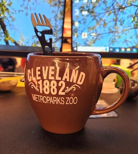 A brown mug - wider at the top than at the base - with the words 
CLEVELAND
1882
METROPARKS ZOO

There is a metal spork sticking out of it.  Well, the knork (knife/fork) half, at least.  There are holes for assorted sizes of hex nuts, as well as a bottle opener, and some large circular hole that I'm not really sure its purpose.  

It is sitting on my desk.  Behind it, softly out of focus, are my two monitors with bckgrounds of an oak tree canopy in early spring - the leaves are tiny and green, the sky is bright blue.  There are also assorted items on the desk, such as a polished sphere of agate, a translucent tea strainer in the shape of a cute little astronaut (or perhaps a deep-sea diver with an old-fashioned diving helmet?), a squishy green stress relief toy in the shape of a tardigrade, and a 5x7 inch plastic magnifier.