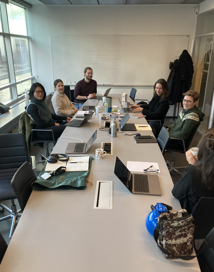 A long grey table around which sit six people on black swivel chairs. Each has a laptop and the table has snacks on it. There is a whiteboard and a window on the left.