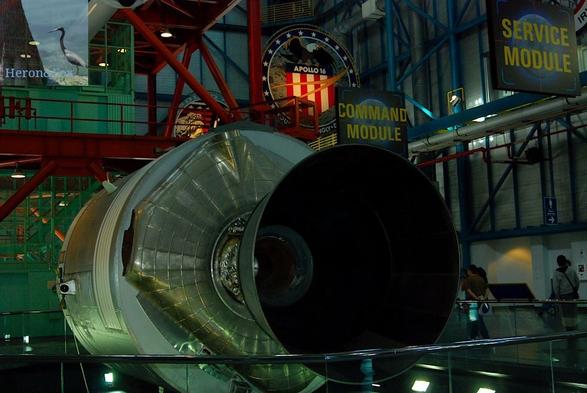 An indoor photograph of a bell-shaped rocket engine nozzle mounted on a gray metal cylinder. Beyond that is a red truss structure. On the right side are two signs reading “Command Module” and “Service Module.”