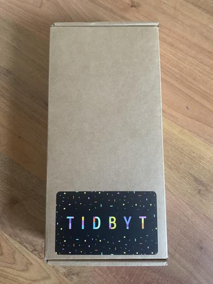 A mysterious cardboard box, with a colourful “Tidbyt” sticker at the bottom! The sticker has a black background, with colourful confetti-like rectangles decorating it.