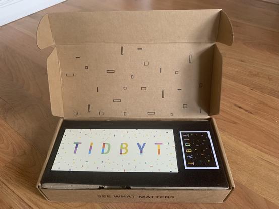 Opening the box reveals another “Tidbyt" sticker, this one still on its’ backing, ready for placement on a laptop. There is also a much larger piece of cardboard, with the Tidbyt logo and a white background. Both are sitting atop a piece of black foam.