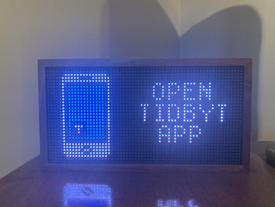 The display is now plugged into power and reads “Open Tidbyt App”, with a pixelated photo of a phone next to it.