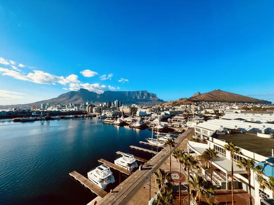 Scenic view of a coastal city with a prominent flat-topped mountain in the background, marina with boats in the foreground, and clear blue skies.
