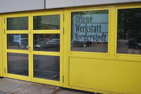 Eingang zur Offenen Werkstatt Norderstedt. Glastür mit gelben Rahmen. Schild mit Schriftzug Offene Werkstatt Norderstedt hängt daneben im Fenster. Die Wand und der Fensterrahmen sind ebenfalls gelb.