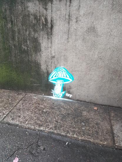 petit champignon bleu turquoise fluo et blanc d'environ 30 cm de haut peint en bas d'un mur, qui regarde le passant avec deux grands yeux