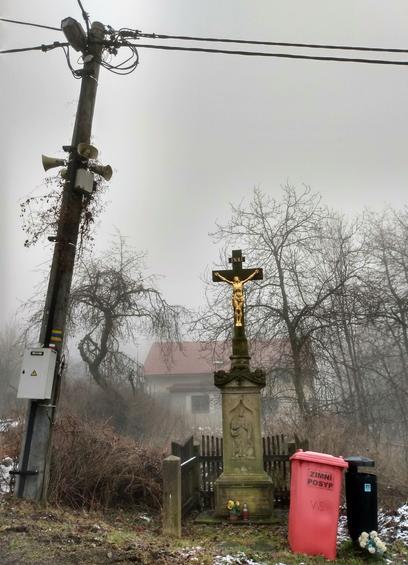 The picture shows different things. A golden Jesus  monument with small yellow flowers in a vase in front of it. A big red plastic rubbish bin and a smaller black one on the right. An old electrical power pole with four plastic speakers attached to it in the upper part on the left. A foggy background with a house and trees in a garden.