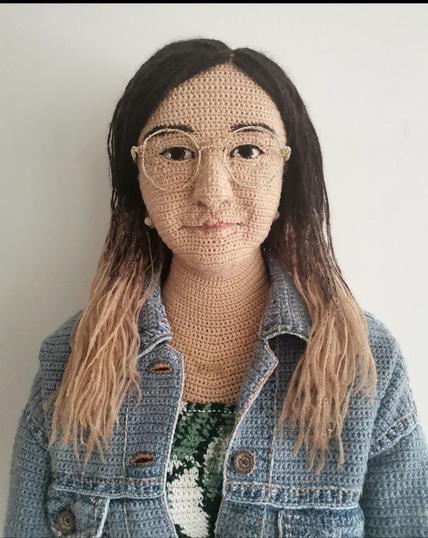 Photography and Art. A color photo
of a lifelike-looking female doll made of knitted or crocheted wool. At first glance, the photograph appears to depict a young woman with long blond, braided hair (slightly darker at the top). She has black eyes, wears round golden glasses, has a medium-sized nose and pink lips. She is dressed in a denim jacket and a white and green woolen sweater. In reality, she is made entirely of wool. A life-size sculpture that can also stand and is completely knitted/crochet. A work of art in which even the buttons and a gold chain are made of wool. Fantastic.