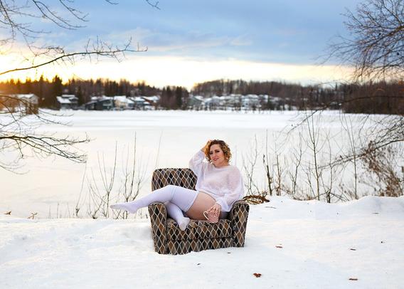 A beautiful woman holds a mug of hot cocoa while stretching her legs across the arm of an easy chair. The setting is a frozen hill overlooking an icy lake in Alaska, with the orange sun creeping up over the trees in the background.