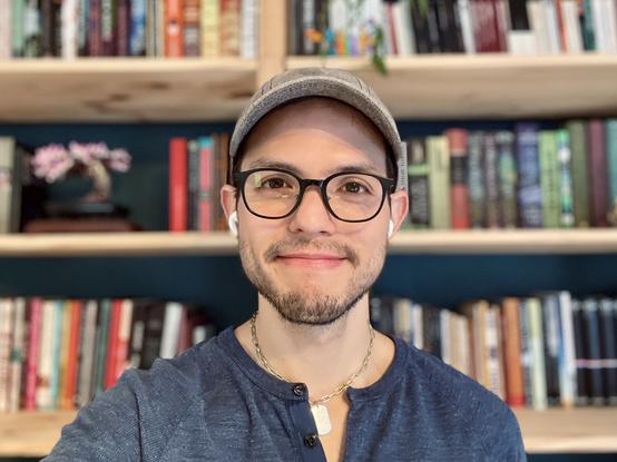 Selfie in front of my bookcase. I’m wearing a blue shirt with the top buttons open, a dog tag necklace can be seen on my neck. I’m wearing my glasses and grey cap.