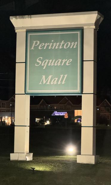 Pre-dawn view of a very large sign in the corner of a shopping center, illuminated by spotlights on the ground front and back that stands like a segmented tan sandstone doorway, over 30 feet tall. It has a rectangular—repeat: *rectangular*— blue-green insert with a thin tan border filling the top half of the arch like the upper panel of a so-called dutch farmdoor. The sign reads in italic, tan letters “Perinton  Square Mall.” Repeat: “Perinton *Square* Mall.”