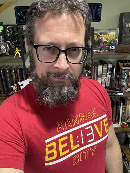 A man with glasses and a salt-and-pepper beard, wearing a red Kansas City Chiefs T-shirt, taking a selfie in front of a bookshelf filled with various decorative items and books.