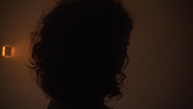 Bild zeigt den Kopf einer Person mit lockigem Haar von hinten vor diffusem Hintergrund. Am linken Bildrand ist ein Scheinwerfer zu sehen.