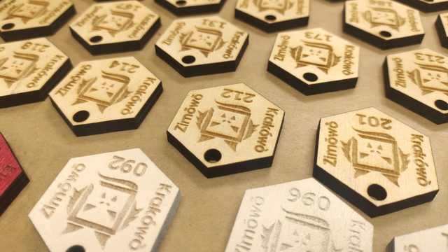 A close-up shot of one of the "ordinary" badges, visible upside down. The inscription "Krakówò zimówò" is noticeable (meaning "Krakow in winter," with the "ó and ò" having a diacritic marks that resembles eyebrows in the emoticon 'ówò', representing a face). Additionally, the sequential numbers on the badges are visible: 201, 212, 214. Also in the frame are fragments of a red badge and pieces of white badges. In the remaining corner of the hexagon, there's a small hole for string attachment. Between the number and the inscription, there's a logo resembling a dragon (a reference to the Wawel Dragon).