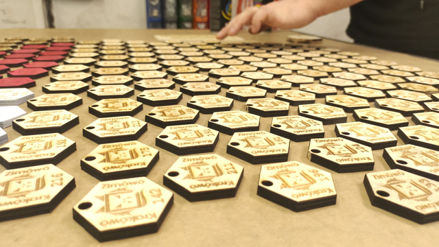 Another perspective of the laser-cut badges, captured at a sharper angle. In the background, containers hanging on the workshop wall and the edge of a paper painting mat, used to protect the table surface from paint stains, are visible.