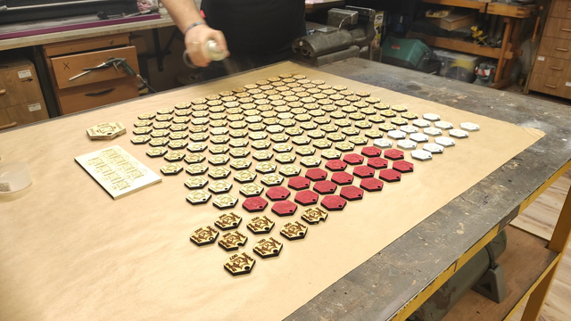 A wide-angle view showcasing the entirety of the table along with the protective paper mat and the rest of the workshop area in the hackerspace. Visible are cabinets, workbenches, a portion of a lathe, a carpentry table, a vice, and the welding argon cylinder positioned under the table. Additionally, a fragment of a figure spraying badges with spray lacquer is visible.