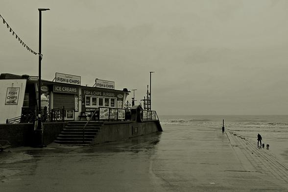 Prom in heavy rain, concrete steps and man walking two dogs, shore break, closed ice cream and fish and chips stores, black and white and gray